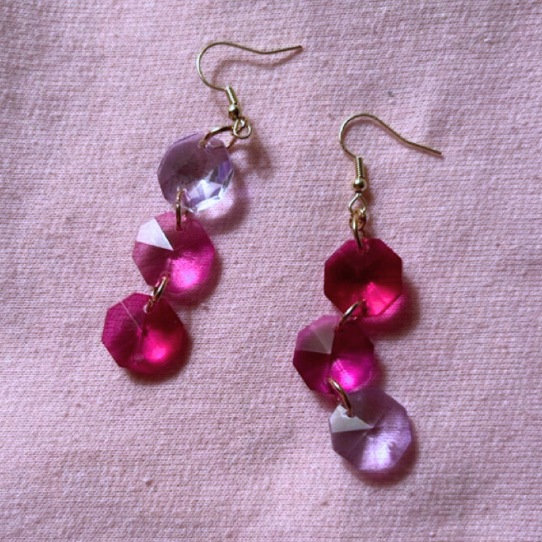 Dangling faceted octagon glass bead earrings. 3 shades of pink beads and gold color fishhook earwires on a light pink fabric background.