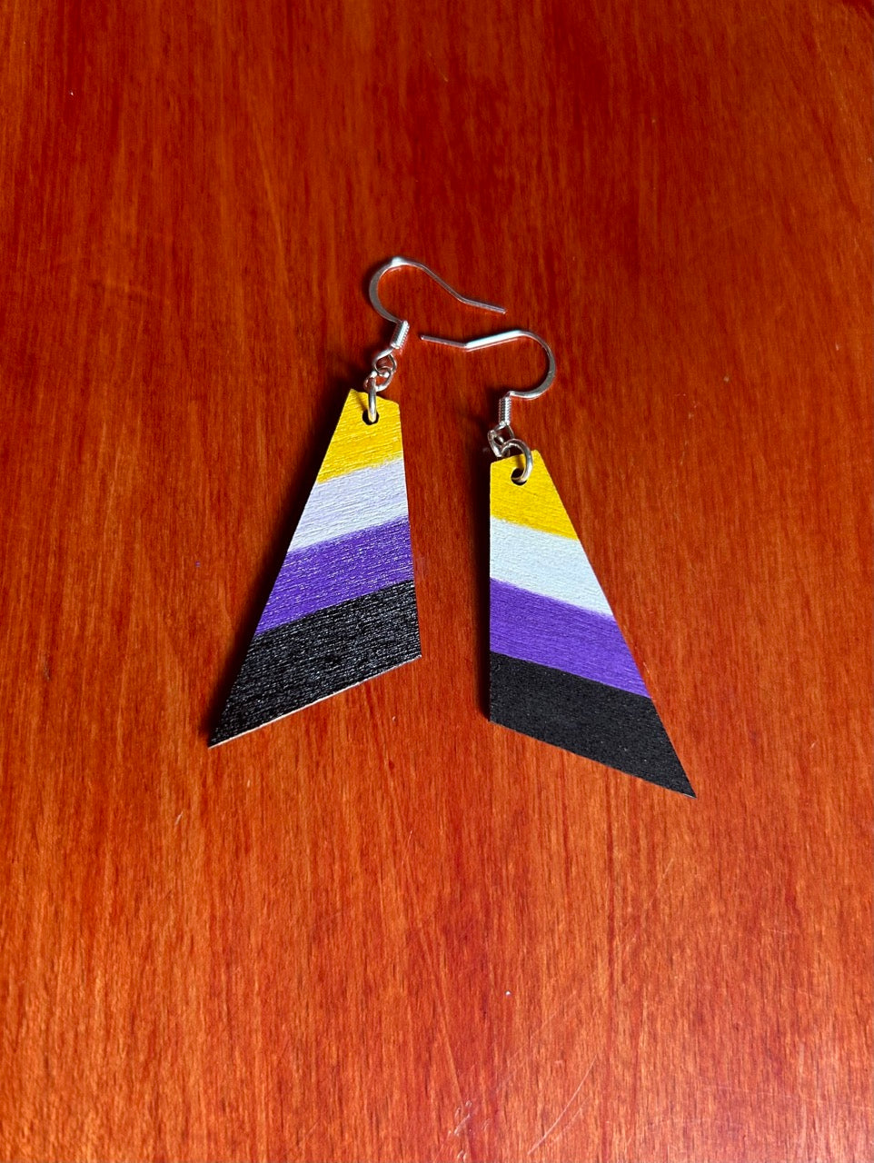 A pair of geometric dangle earrings with angled horizontal stripes in yellow, white, purple, and black, hanging from silver fishhook wires on a red-brown wooden surface.