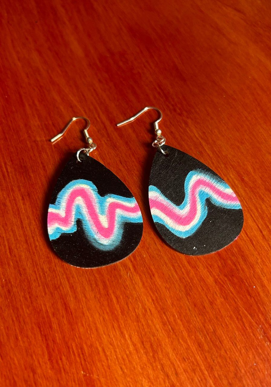 Back side of a pair of teardrop-shaped wooden dangle earrings with a black base and wavy pink, blue, and white stripes. Includes silver fishhook wires on a red-brown wooden surface.