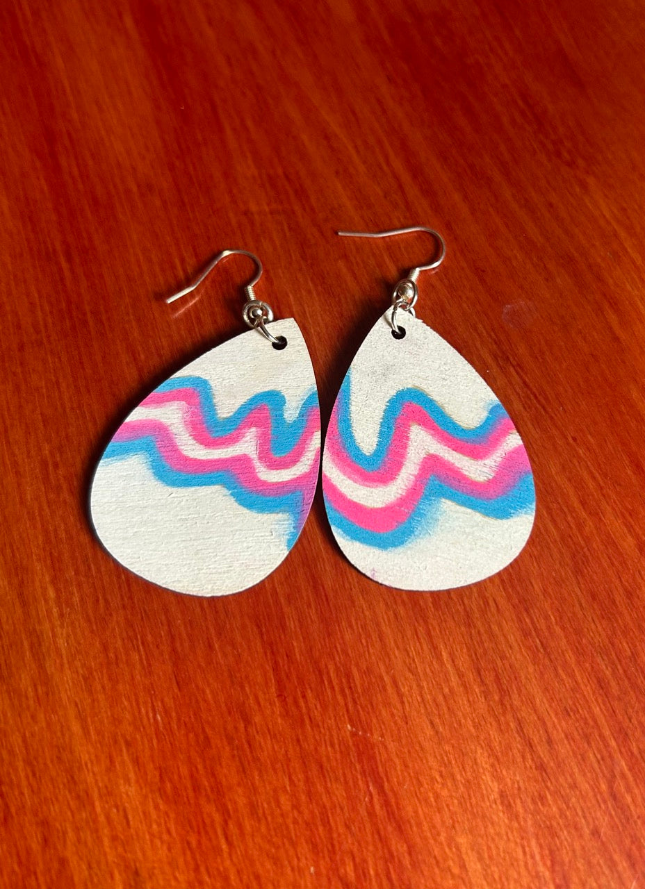 Front side of a pair of teardrop-shaped wooden dangle earrings with a white base and wavy pink and blue stripes. Includes silver fishhook wires on a red-brown wooden surface.