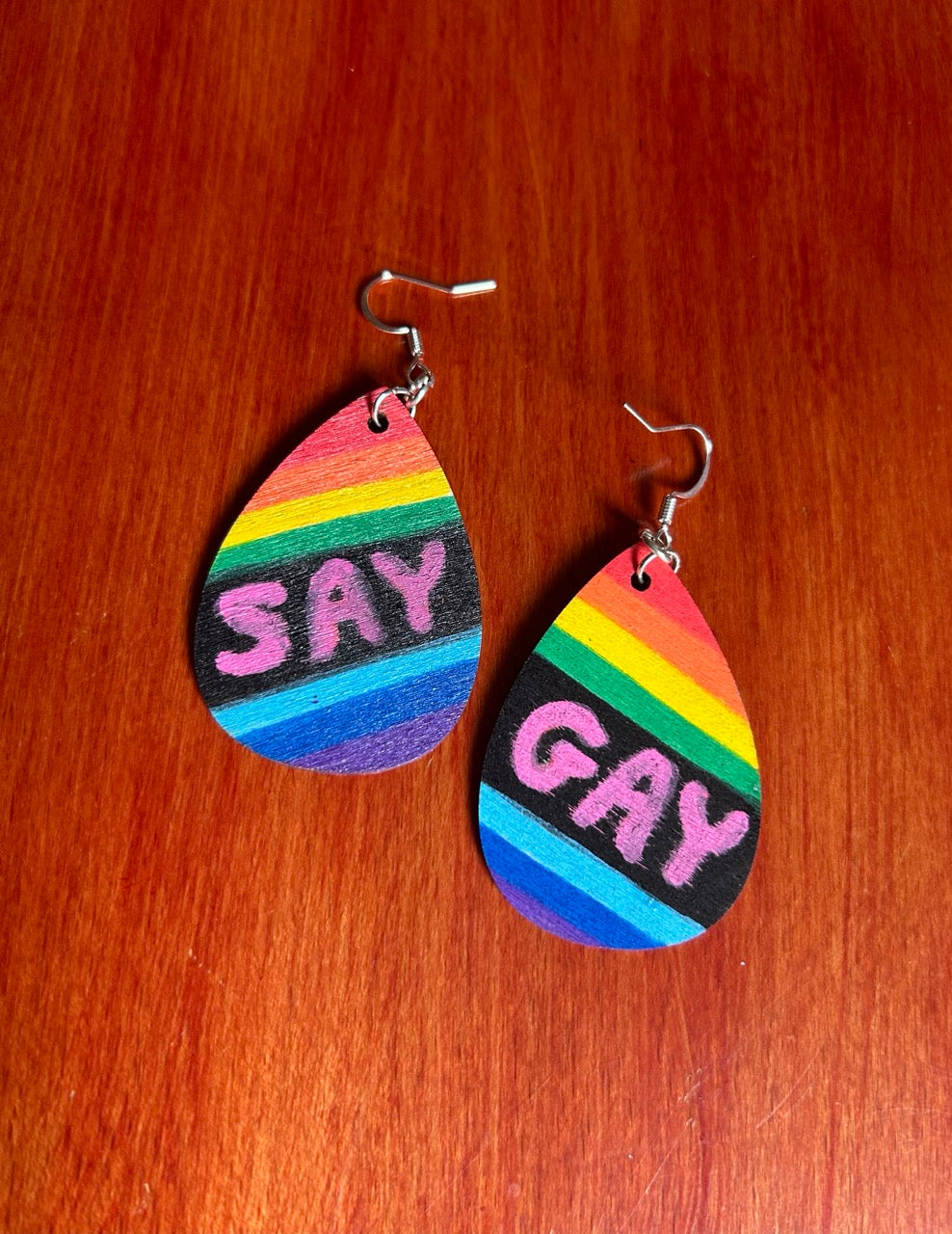 Front side of teardrop-shaped wooden dangle earrings with rainbow stripes and the words 'SAY GAY' in pink, displayed on a red-brown wooden surface.