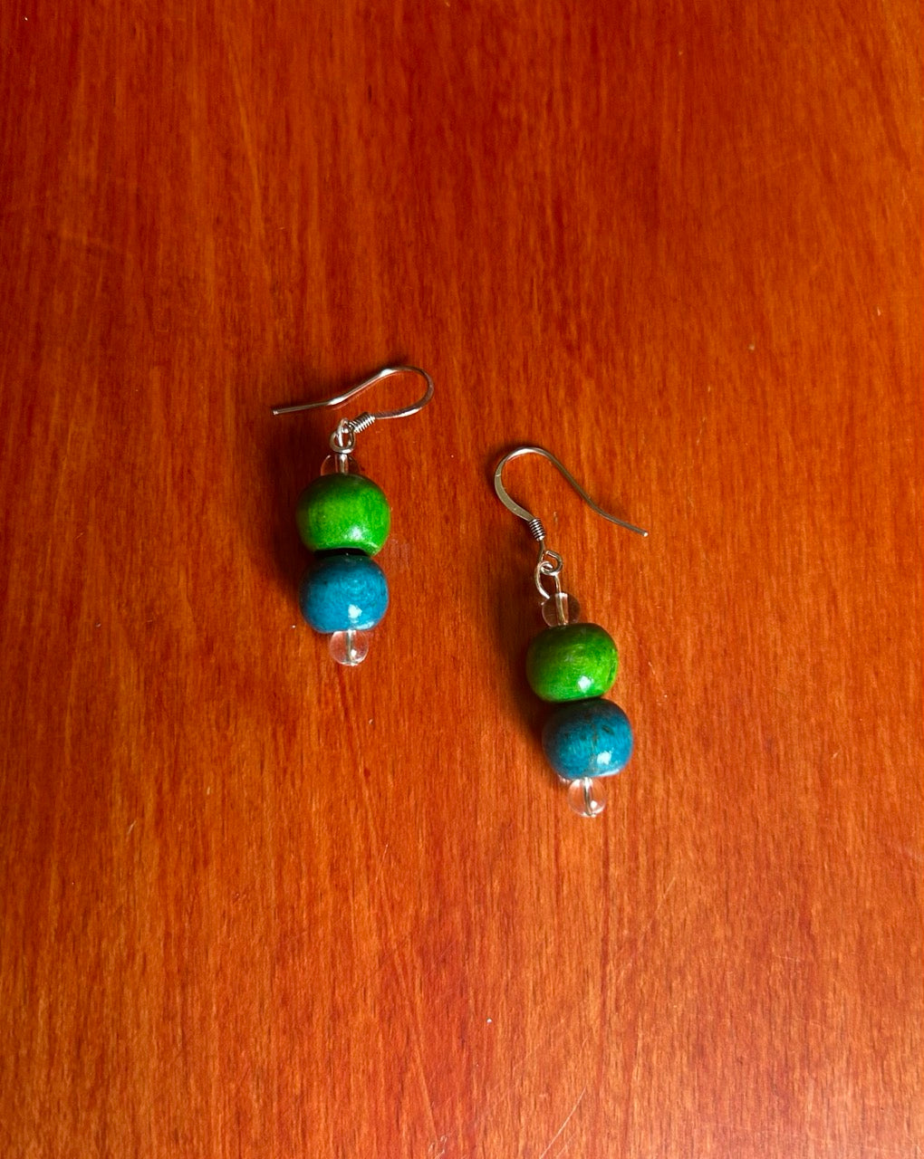 A pair of handmade beaded dangle earrings with green and blue wooden beads on stainless steel fishhook wires, resting on a red-brown wooden surface.