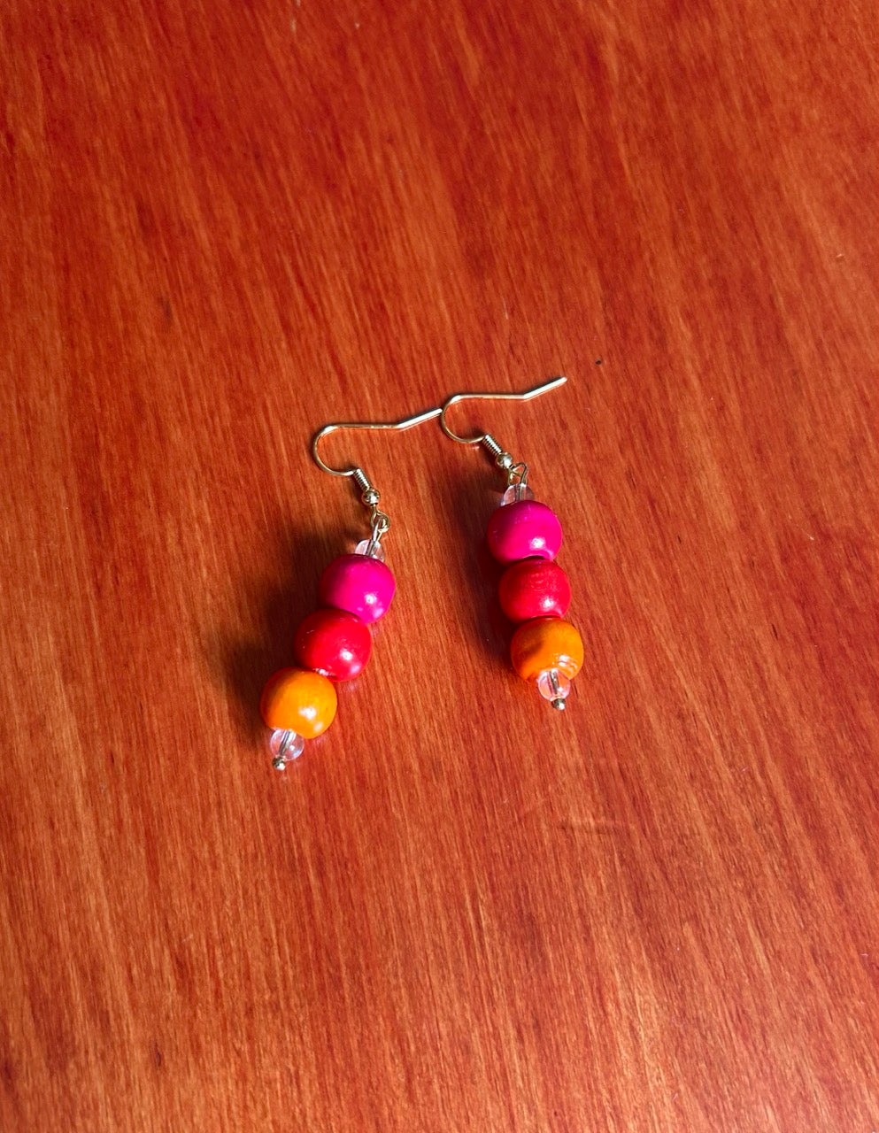 A pair of handmade beaded dangle earrings with pink, red, and orange wooden beads on silver fishhook wires, resting on a red-brown wooden surface.
