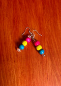 A pair of handmade beaded dangle earrings with pink, yellow, and blue wooden beads on silver fishhook wires, resting on a red-brown wooden surface.