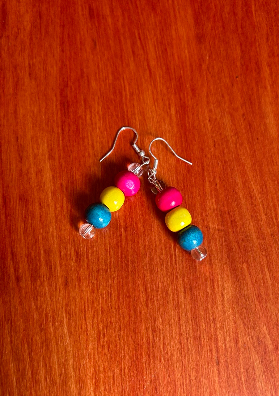 A pair of handmade beaded dangle earrings with pink, yellow, and blue wooden beads on silver fishhook wires, resting on a red-brown wooden surface.