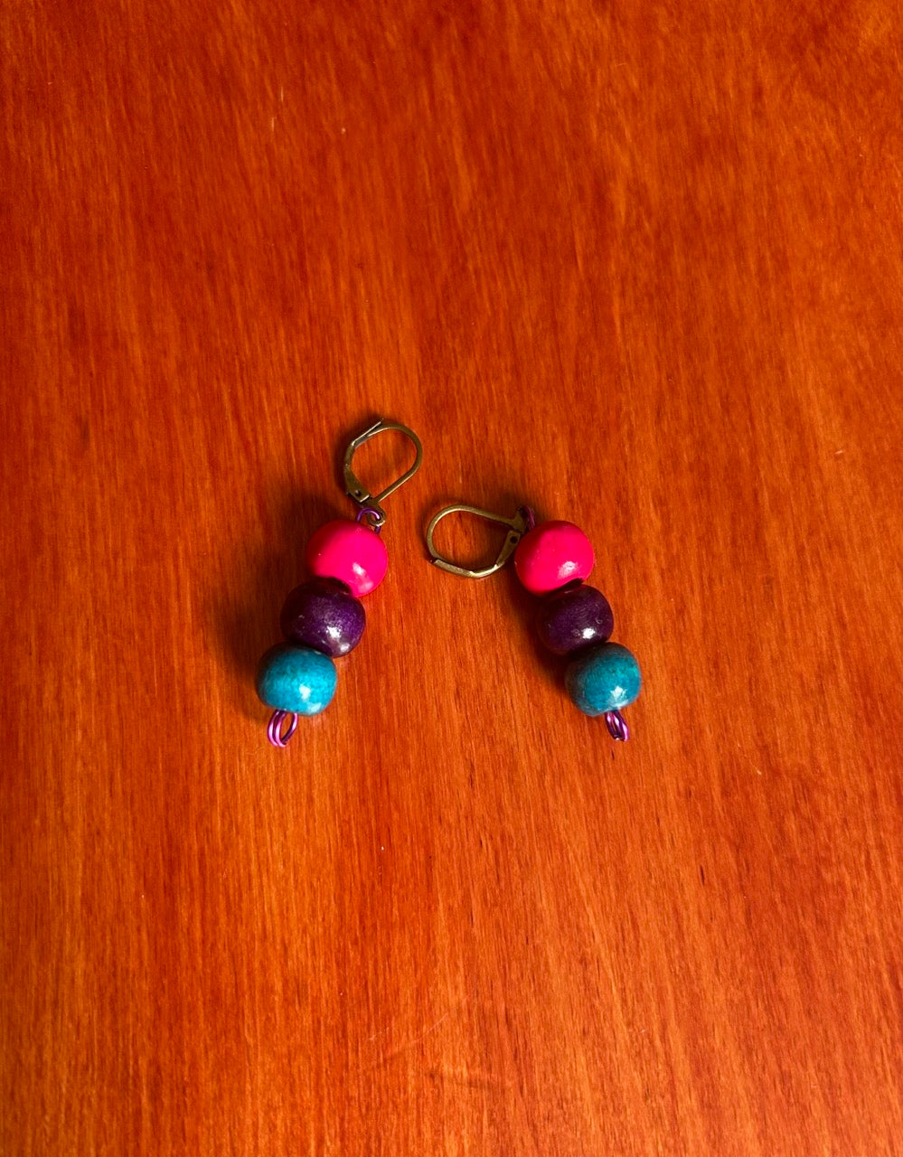 A pair of handmade beaded dangle earrings with pink, purple, and blue wooden beads on bronze clasp wires, resting on a red-brown wooden surface.