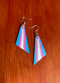 A pair of handmade geometric dangle earrings with a pink, white, and blue vertical striped pattern, hanging from silver fishhook wires on a red-brown wooden surface.