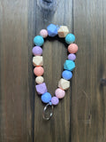 A pastel-colored beaded keychain with geometric and round beads in shades of blue, pink, purple, and peach. It is attached to a silver metal key ring. Displayed on a dark wood background.