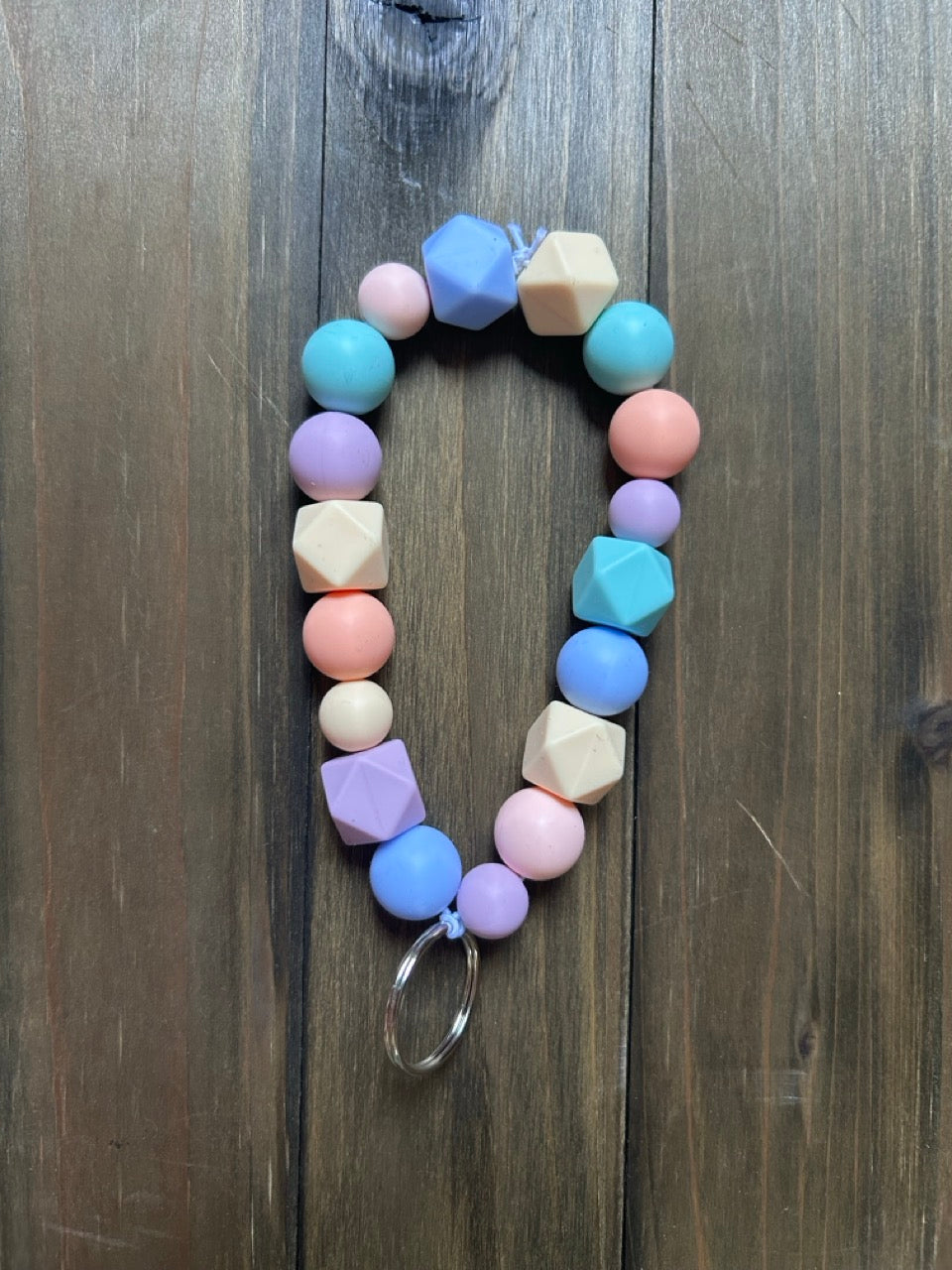 A pastel-colored beaded keychain with geometric and round beads in shades of blue, pink, purple, and peach. It is attached to a silver metal key ring. Displayed on a dark wood background.