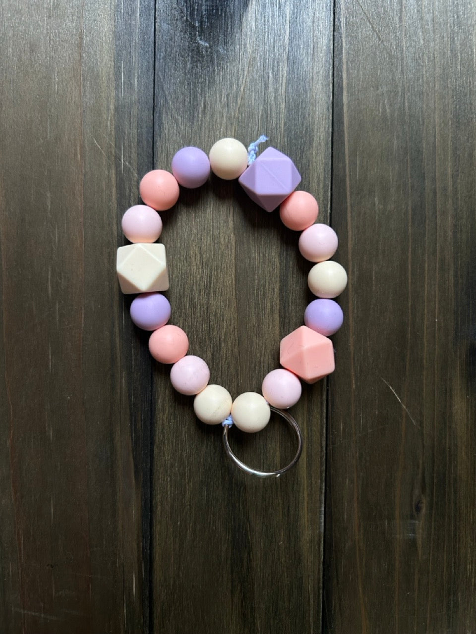 A pastel beaded keychain with round and faceted beads in pink, lavender, cream, and peach, attached to a silver metal ring, displayed on a dark wood background.