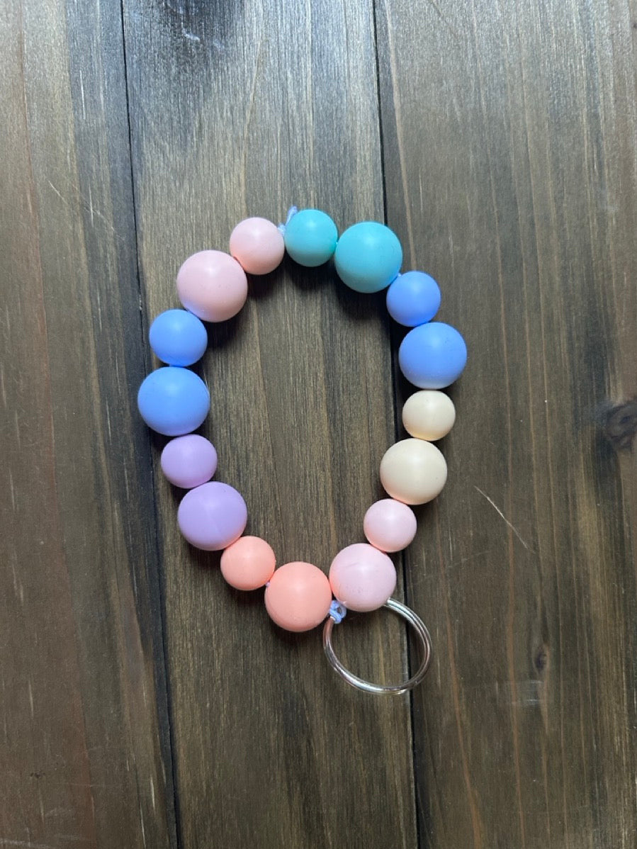 A beaded keychain with pastel-colored rainbow round silicone beads. It is attached to a silver metal split ring. Displayed on a dark wood background.