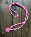 A pink-to-white gradient polymer clay bead lanyard with a flower-shaped pink focal bead above a silver key ring. Displayed on a dark wood background.