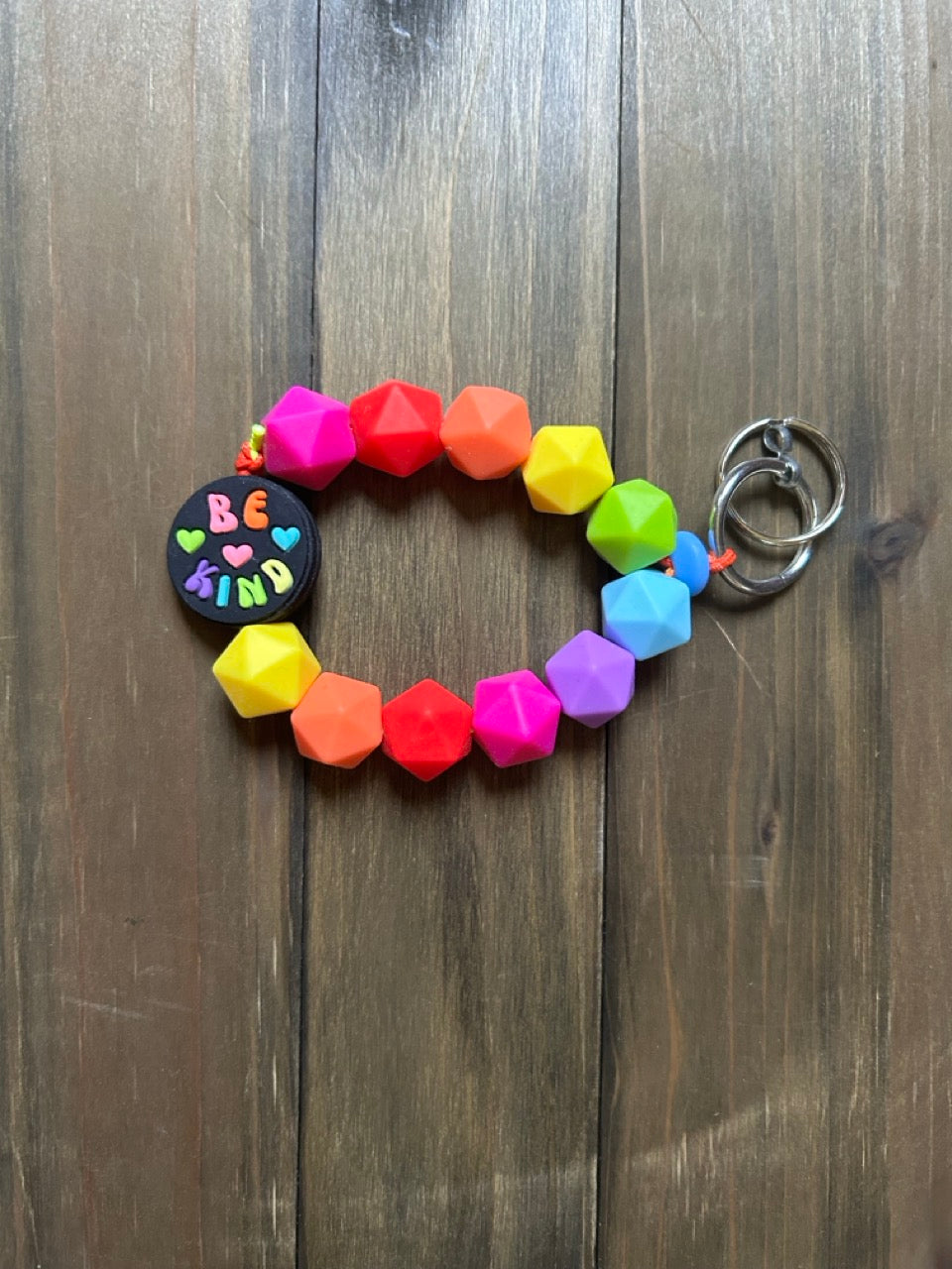 A brightly-colored silicone beaded keychain; includes faceted geometric beads in rainbow order. Features a round black focal bead  that says 'BE KIND' in multicolored letters. shown on a dark wood background.