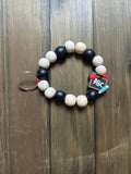 A beaded keychain featuring alternating matte black and natural light wood beads. In the middle of the keychain is a small school-themed charm shaped like a chalkboard with ABC on it, as well as a red apple and pencil. Displayed on a dark wood background.