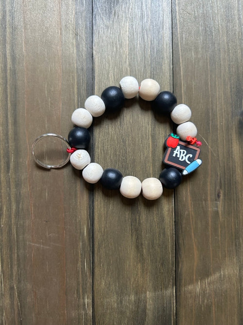 A beaded keychain featuring alternating matte black and natural light wood beads. In the middle of the keychain is a small school-themed charm shaped like a chalkboard with ABC on it, as well as a red apple and pencil. Displayed on a dark wood background.