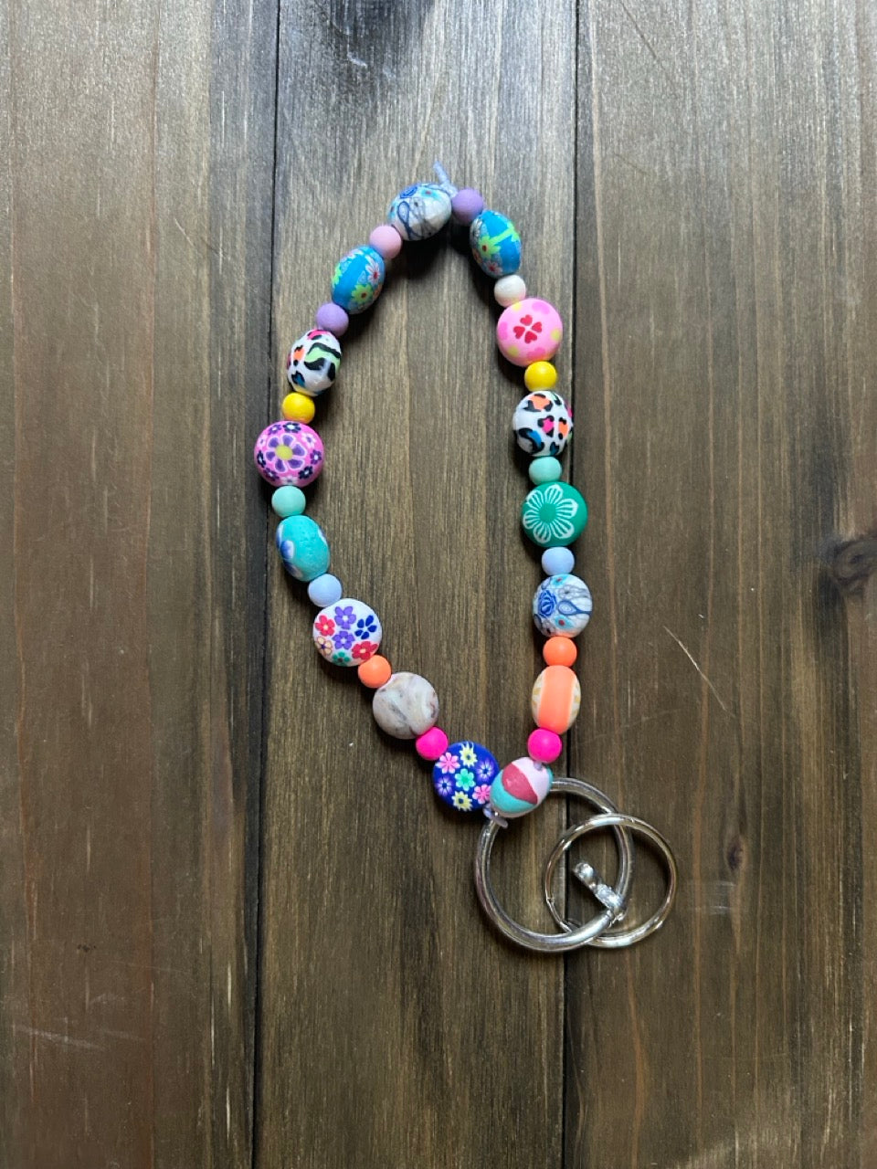 A colorful beaded keychain with patterned and solid round beads in pink, blue, purple, orange, and green, attached to two silver metal rings, displayed on a dark wood background.