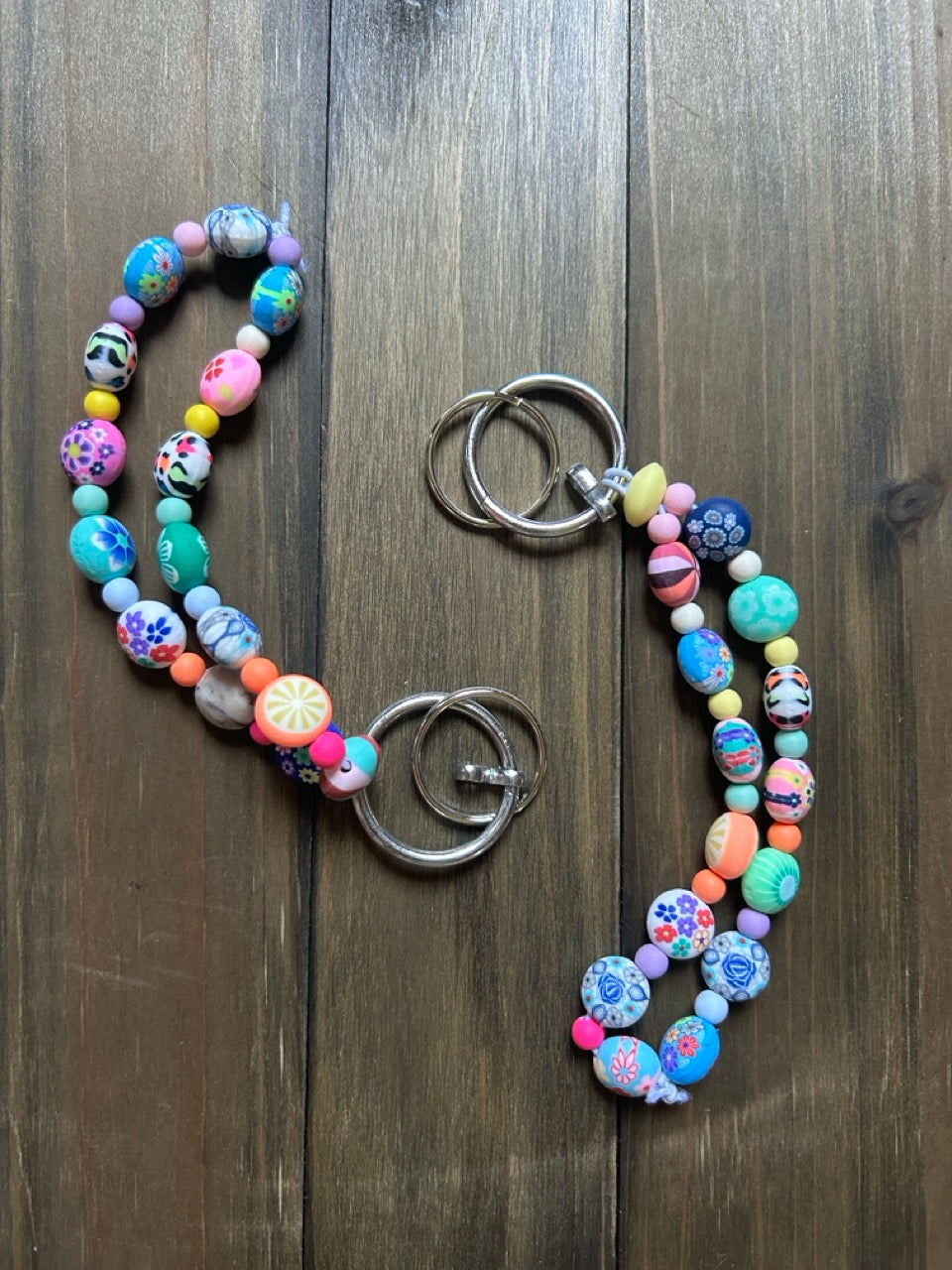 Two beaded keychains with two silver metal key rings, featuring colorful patterned and solid beads in pink, blue, green, yellow, and orange, laid flat on a dark wood background.