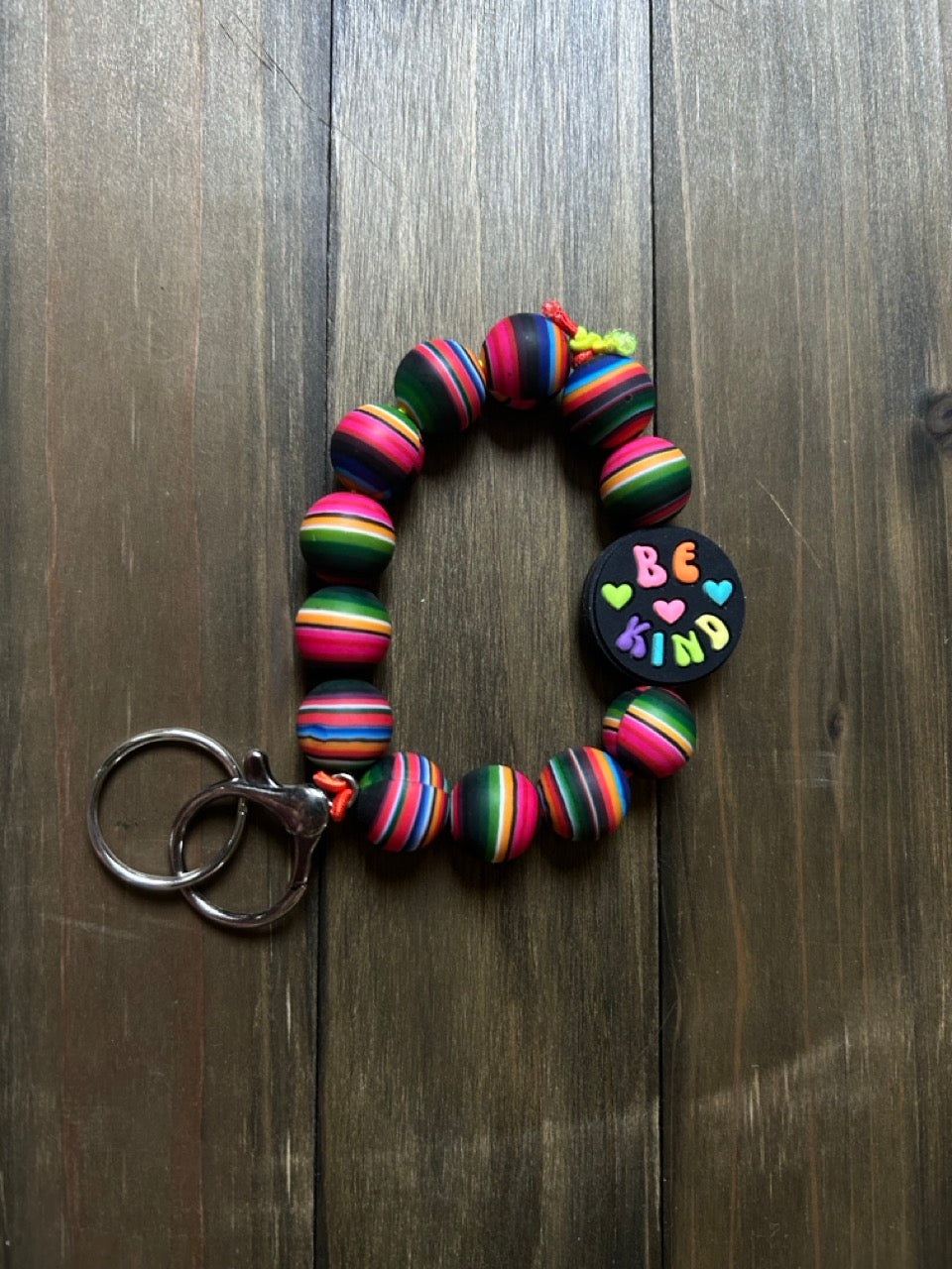 A silicone beaded wristlet with colorful striped beads and a round black charm that says 'BE KIND' in multicolored letters. The wristlet is resting on a dark wood background.
