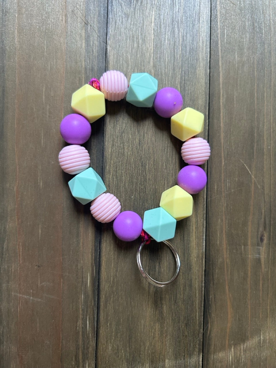A pastel-colored beaded keychain with purple, pink, light yellow, and mint green geometric silicone beads, resting on a dark wood background.