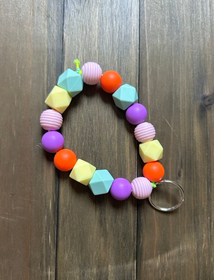 A colorful beaded keychain with geometric silicone beads. The beads are colored in pastel mint, purple, pink, bright orange, and pale yellow. It features a silver key ring,. Displayed on a dark wood background.