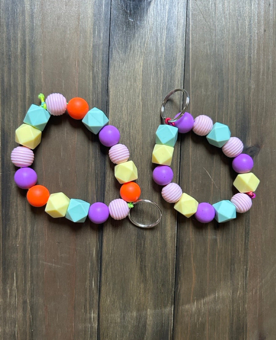 Two colorful beaded keychains with geometric silicone beads in purple, yellow, pink, orange, and light blue, displayed on a dark wood background.