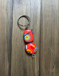 A handmade keychain featuring two red millefiori glass beads with colorful abstract floral patterns. It is attached to a silver metal split ring, and displayed on a dark wood background.