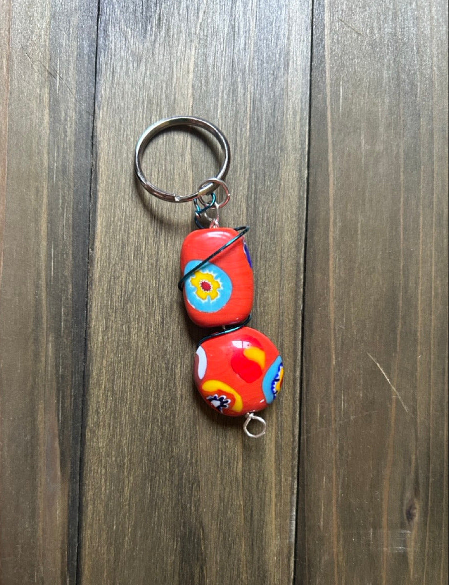 A handmade keychain featuring two red millefiori glass beads with colorful abstract floral patterns. It is attached to a silver metal split ring, and displayed on a dark wood background.