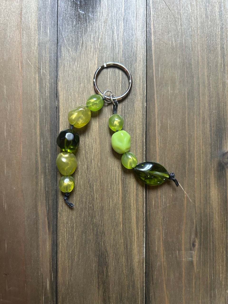 A handmade vintage-bead keychain with translucent beads in multiple shades of green. The beads are attached to a silver split ring. The keychain is displayed on a dark wood background.