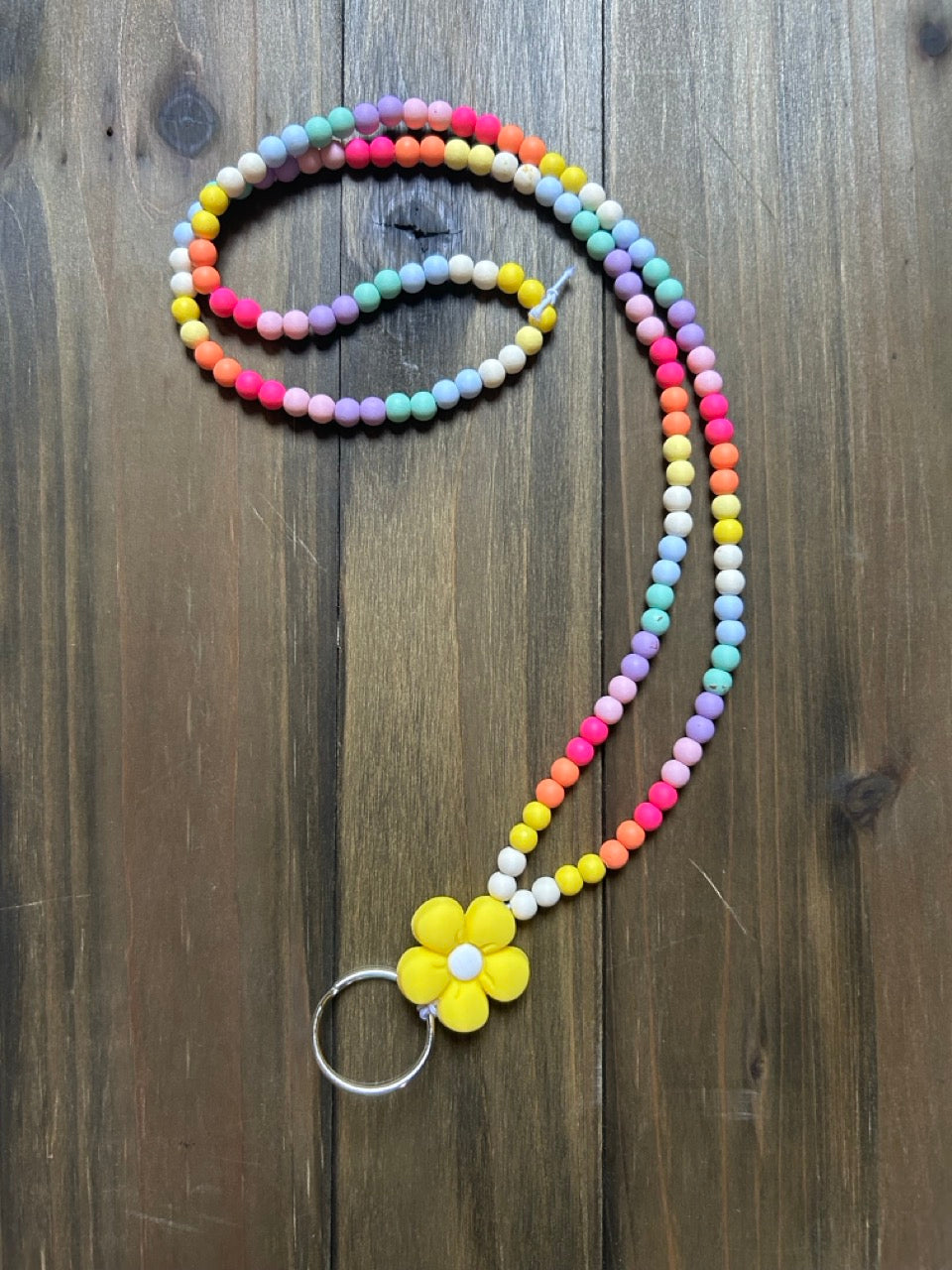 A pastel rainbow gradient beaded lanyard. At the bottom of the lanyard is yellow flower charm and a silver key ring. Displayed on a dark wood background.