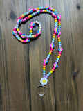 A brightly colored rainbow beaded lanyard. In between each segment of rainbow beads is a with rainbow heart bead. At the base on the lanyard is a white flower smiley face charm above a silver key ring. Displayed on a dark wood background.