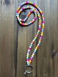 A beaded lanyard with a mix of bright and pastel-colored round beads in light pink, hot pink, yellow, blue, minty green, orange, white, and purple. A silver ring clasp is attached to the base of the lanyard. The lanyard is displayed on a dark wood background.