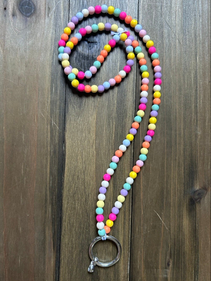 A beaded lanyard with a mix of bright and pastel-colored round beads in light pink, hot pink, yellow, blue, minty green, orange, white, and purple. A silver ring clasp is attached to the base of the lanyard. The lanyard is displayed on a dark wood background.