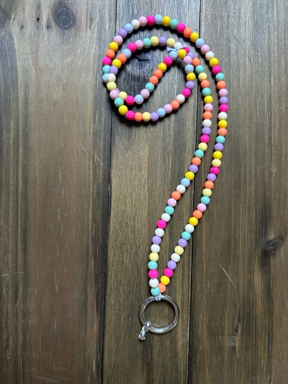 A beaded lanyard with pastel-colored round beads in pink, yellow, blue, green, orange, and purple, featuring a silver-toned split ring clasp, displayed on a wood background.