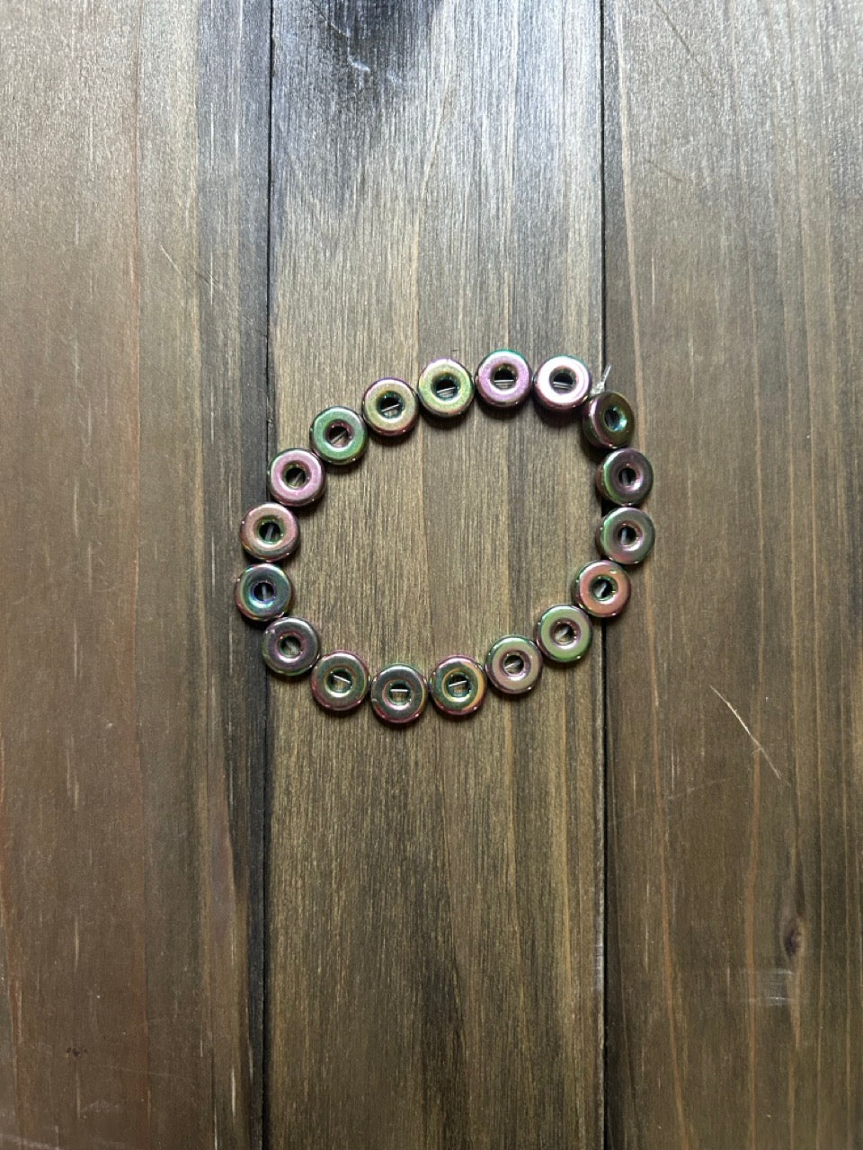 A beaded bracelet made of small iridescent donut-shaped beads in shades of green and pink, on a dark brown wooden surface.