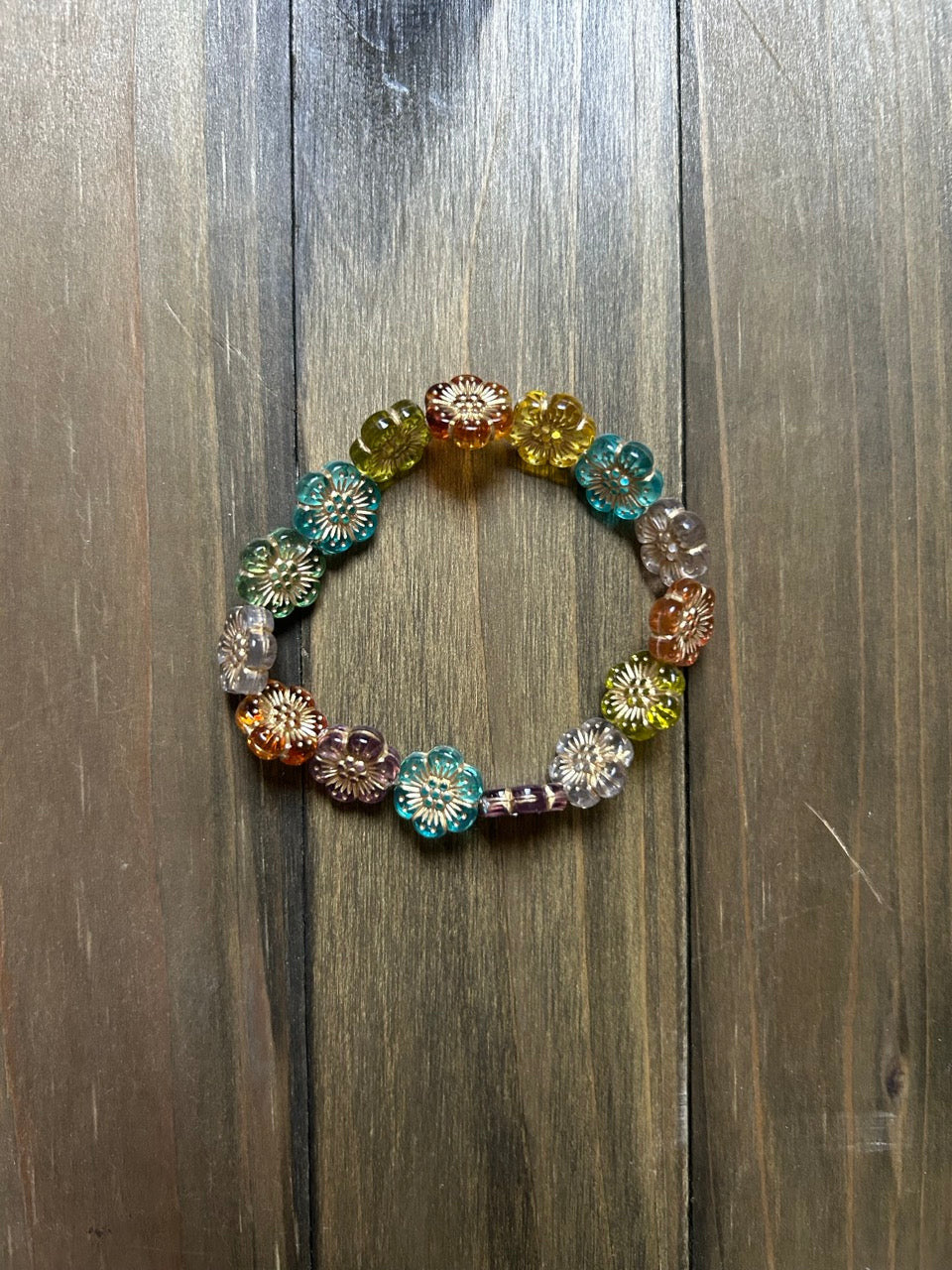 A multicolor beaded stretch bracelet featuring flower-shaped beads in various colors and metallic filigree details, displayed on a dark wood background.