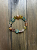 A multicolor beaded stretch bracelet featuring flower-shaped beads in various colors and metallic filigree details, displayed on a dark wood background.