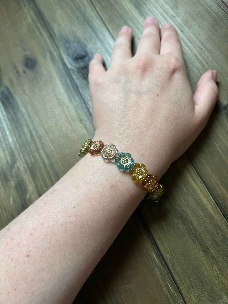 A close-up of a person's wrist wearing a beaded stretch bracelet with colorful floral beads with gold details. The hand is resting on a dark wood surface.