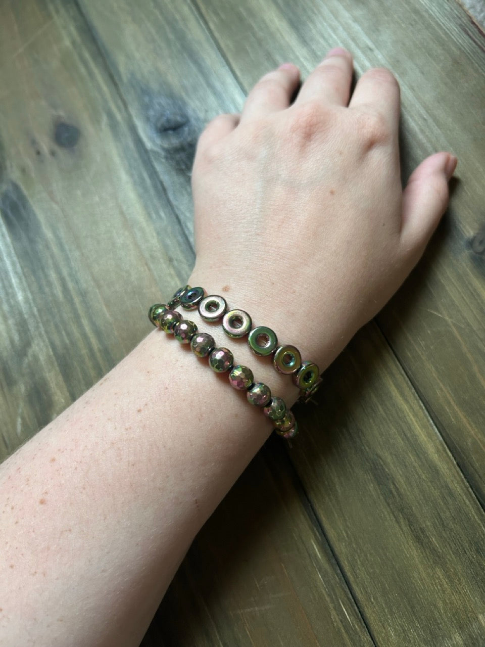 A close-up of a person's wrist wearing two beaded bracelets, resting on a rustic wooden surface. One bracelet features flat metallic donut-like beads, and the other features faceted round beads. Both show multiple tones of muted green and pink.