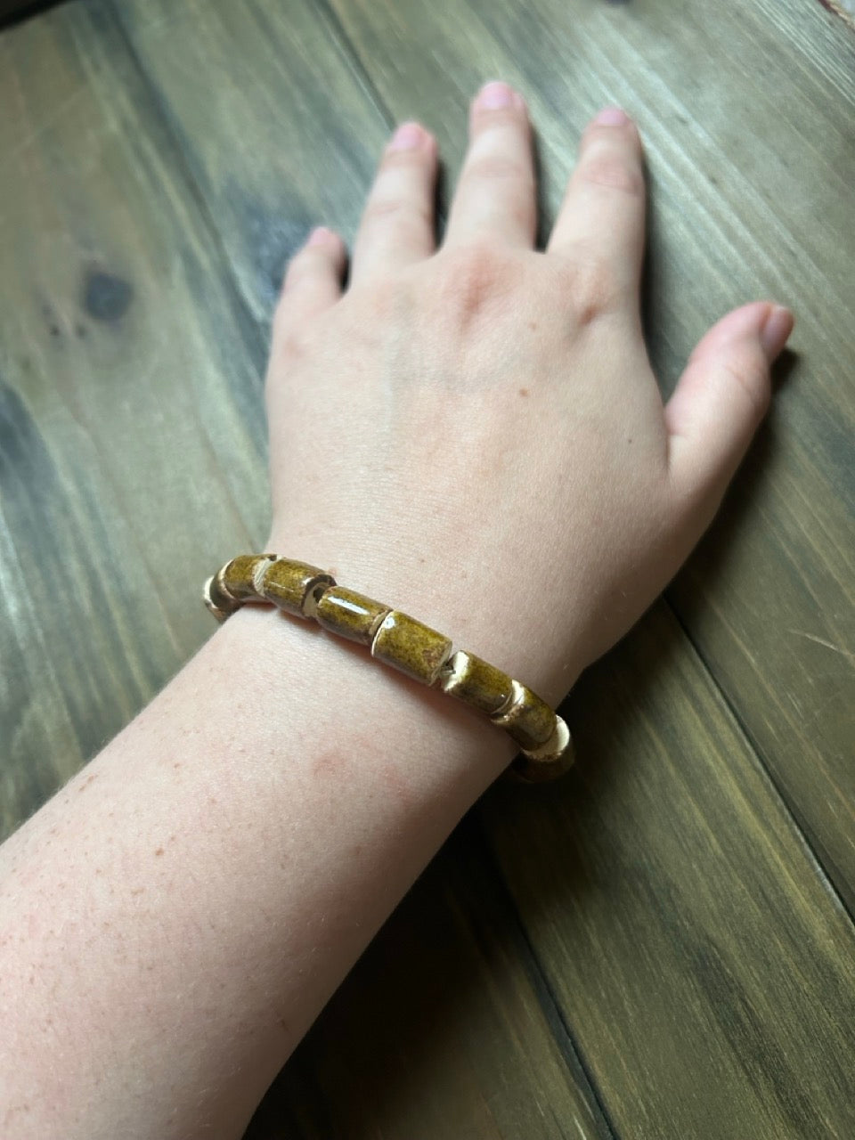A person's wrist wearing a brown ceramic beaded bracelet with cylindrical beads, resting on a dark wood surface.