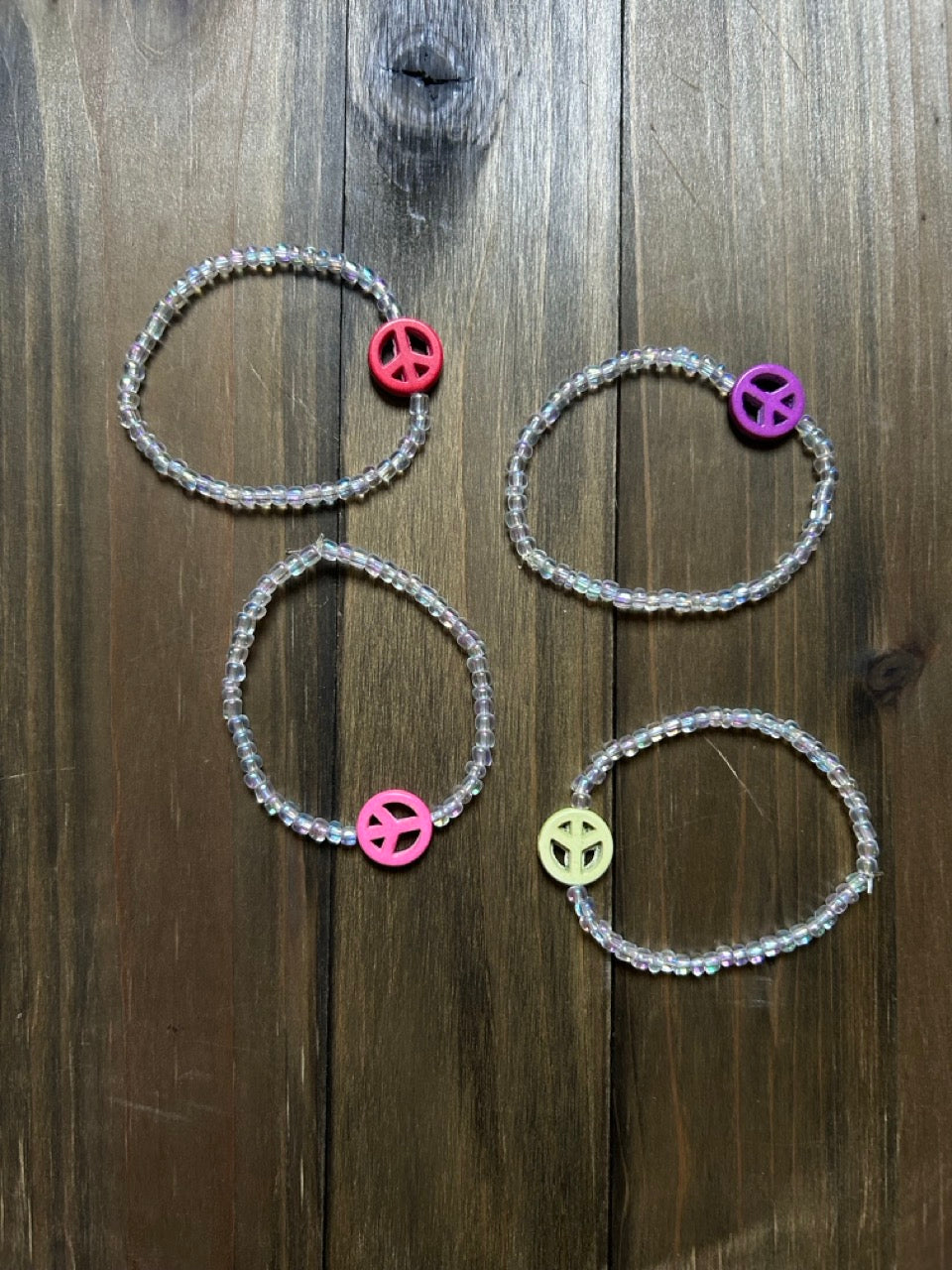Four beaded bracelets with peace sign charms arranged on a dark wood background. The bracelets have clear iridescent beads and feature a central  peace sign bead in pink, purple, light green, and red.