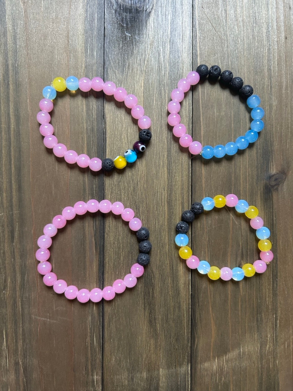 Four glass beaded bracelets arranged on a dark wood background. The bracelets feature pink, light blue, yellow glass beads as well as black lava stone beads. One of the bracelets contains evil eye beads.