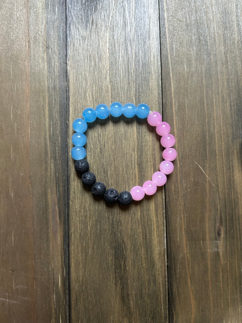 A beaded stretch bracelet with three distinct sections: light blue glass beads,  black lava stone beads, and light pink glass beads, laid flat on a dark wood background.