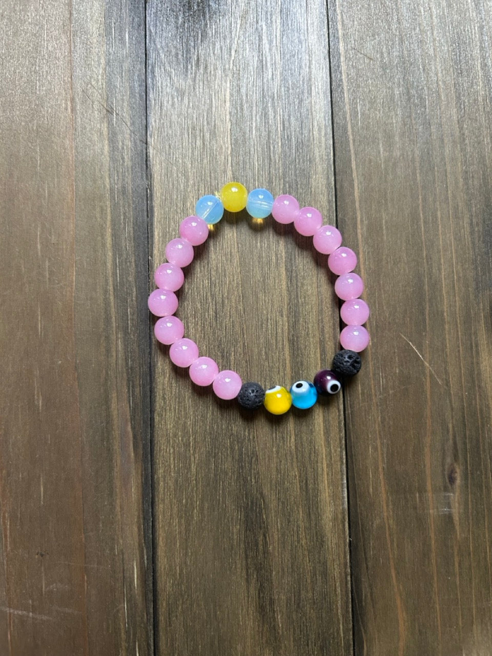 A beaded stretch bracelet with round, light pink, light blue, and yellow glass beads. In between two black lava stone beads on the bracelet is a yellow, a blue, and a dark purple evil eye bead. Displayed on a dark wood background.
