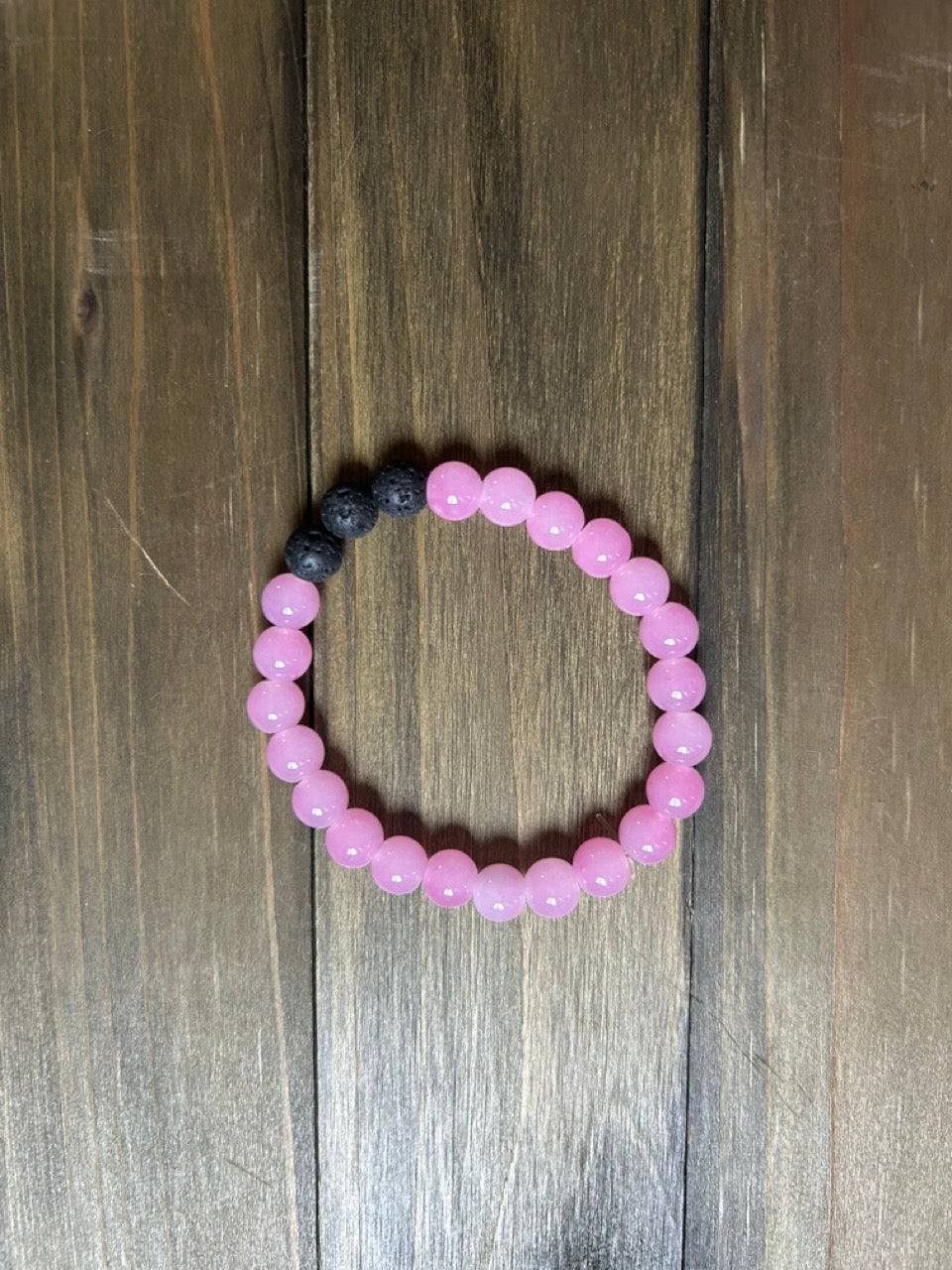 A beaded stretch bracelet made of round, light pink glass beads with a small section of black lava stone beads, displayed on a dark brown wooden surface.
