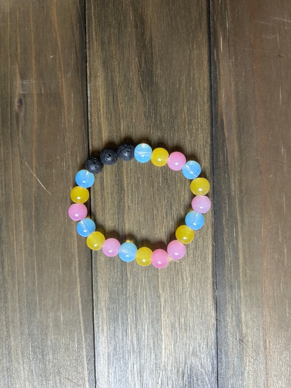 A beaded stretch bracelet with alternating pastel pink, light blue, and yellow beads, plus a small section of black lava stone beads, displayed on a dark brown wooden surface.
