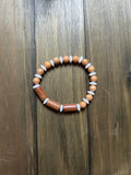 A beaded stretch bracelet made of pale and golden brown wooden beads. Round, pale wooden beads have white spacer bead in between, while the center of the bracelet has golden brown cylindrical wooden beads with white spacer beads in between. Displayed on a dark brown wooden surface.