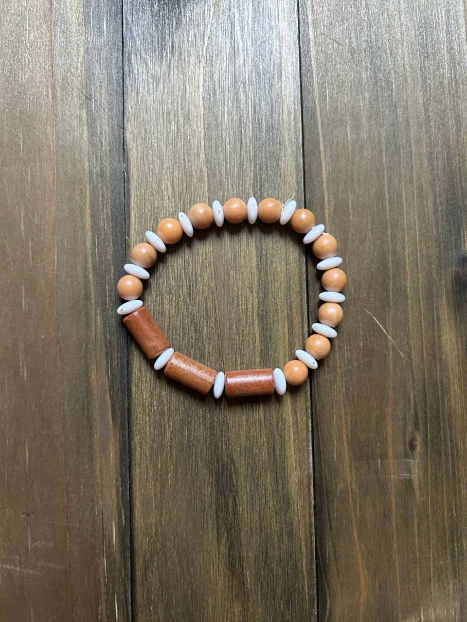 A beaded stretch bracelet made of pale and golden brown wooden beads. Round, pale wooden beads have white spacer bead in between, while the center of the bracelet has golden brown cylindrical wooden beads with white spacer beads in between. Displayed on a dark brown wooden surface.