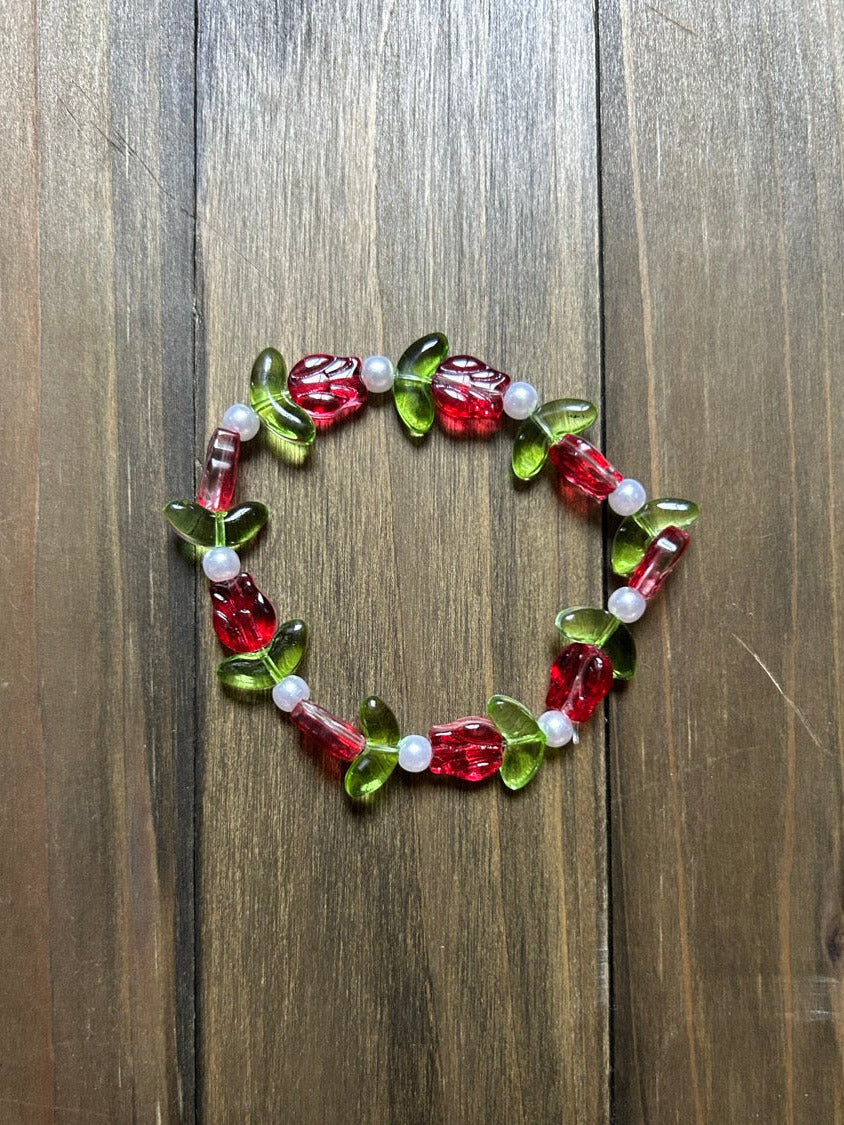 A beaded stretch bracelet featuring red tulip-shaped beads, green leaf-shaped beads, and small white pearl-like beads, displayed on a dark brown wooden surface.