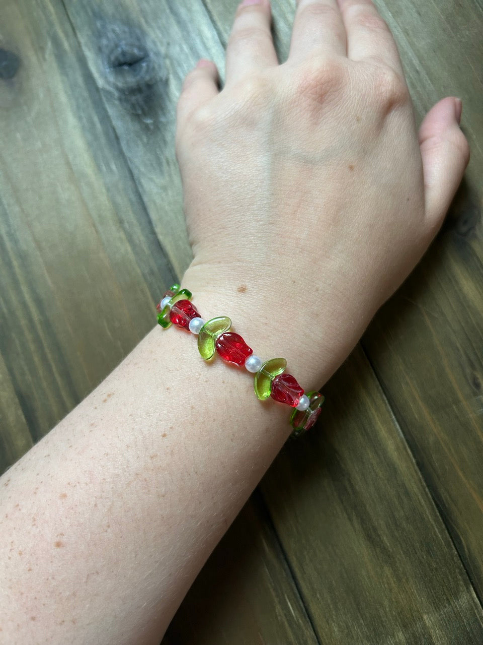 A close-up of a person's wrist wearing a beaded bracelet made of red tulip-shaped beads with green leaves and small white pearl accents, resting on a dark wood background.