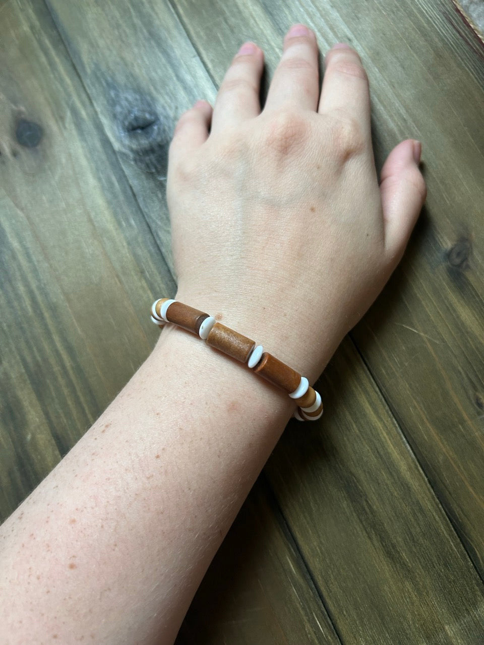 A close-up of a person's wrist wearing a beaded bracelet made of brown wooden beads with white spacer beads. The hand is resting on a dark wood plank surface.